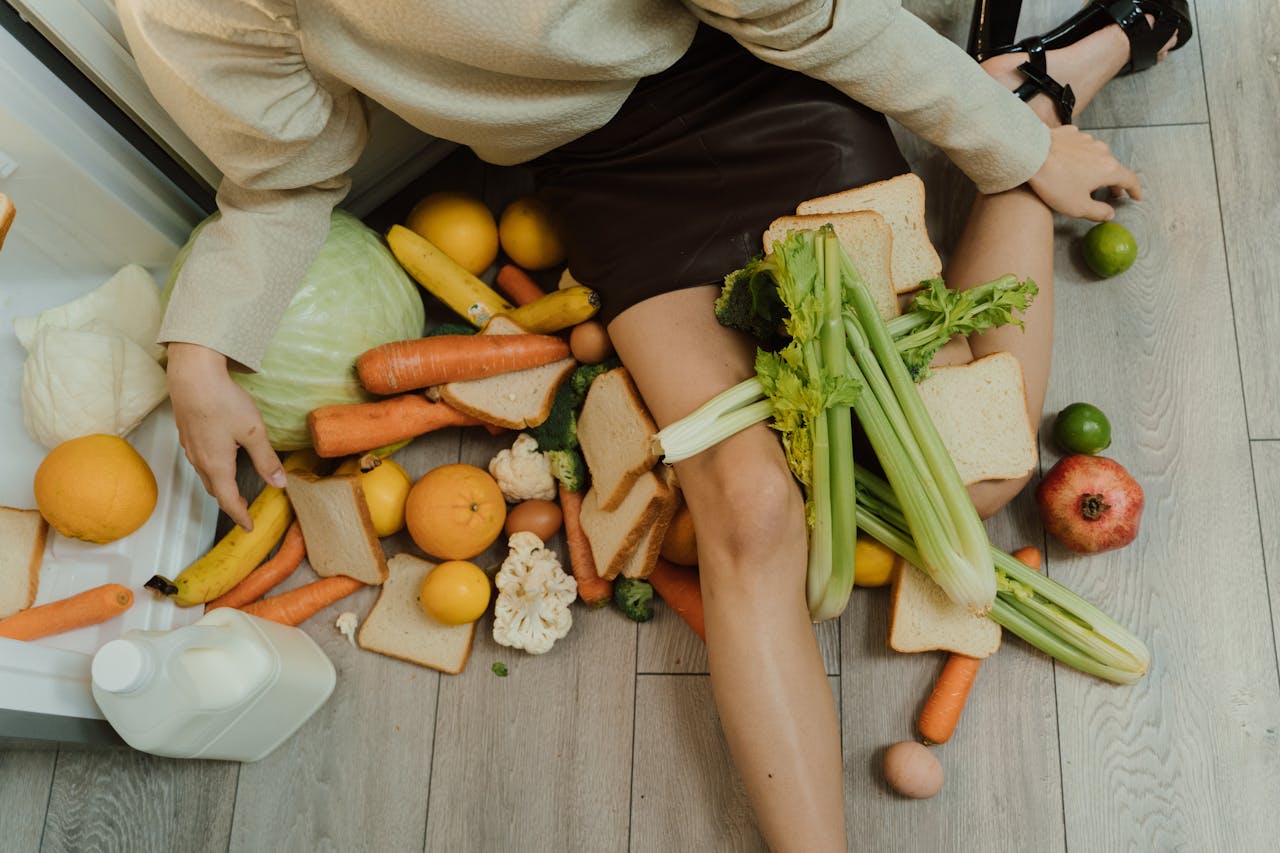 Home A woman amidst spilled groceries, highlighting food waste and consumerism with vegetables, fruits, and bread.